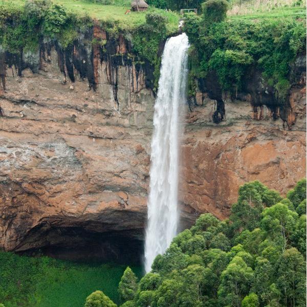 A stunning view of the Sipi Falls waterfall, cascading down rocky cliffs amidst lush green vegetation, with mist rising from the base and the vibrant landscape of Mount Elgon in the background.